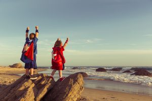 A young boy and girl dressed as a superheroes stands on a rock with arms raised while on a California Beach. The surf and waves wash the sand around them and they are ready to work as a team to accomplish great things as they look out to sea. Image taken in Orange County, California, USA.